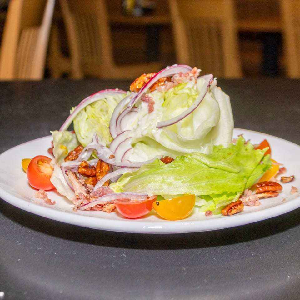 Iceberg wedge salad with red onion, tomatoes, bacon, and pecans on a white plate.