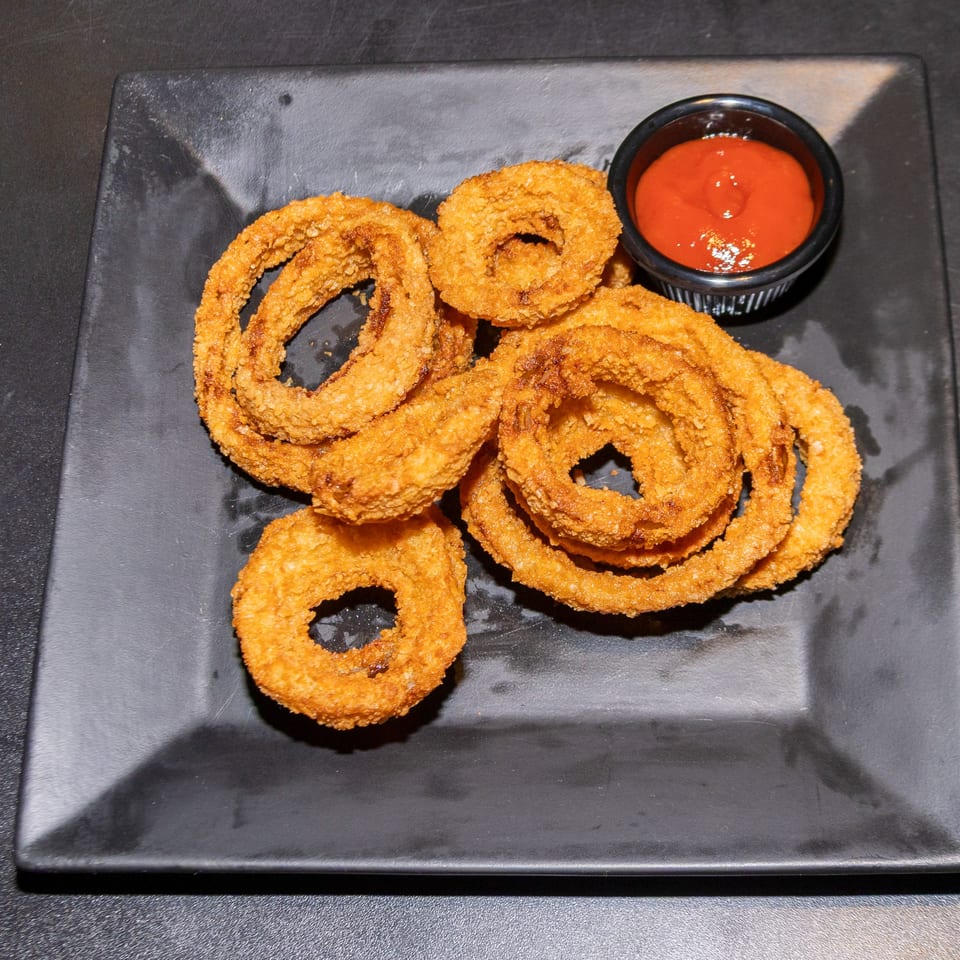 Golden-brown onion rings piled on a black plate with a side of red dipping sauce.