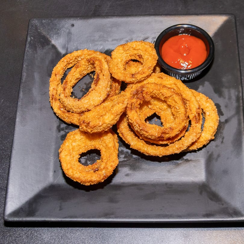 Golden-brown onion rings piled on a black plate with a side of red dipping sauce.